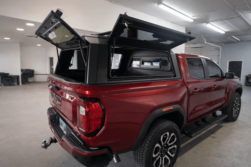 Red GMC Canyon pickup truck with a black aluminum truck cap installed, rear doors open showing interior access, photographed in a garage showroom.