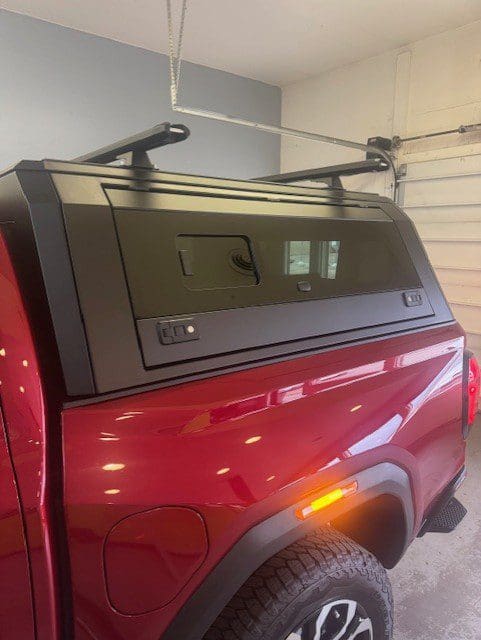 Close-up side view of a matte black truck cap with a sliding side window and roof rack installed on a red pickup truck, shown in a garage.