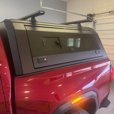 Close-up side view of a matte black truck cap with a sliding side window and roof rack installed on a red pickup truck, shown in a garage.