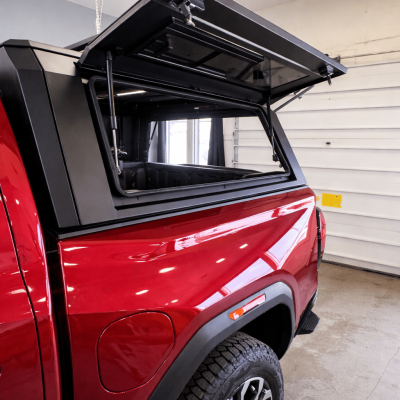 Driver-side view of a red GMC Canyon with a matte black truck cap, side door propped open revealing the enclosed cargo area interior, photographed in a garage.
