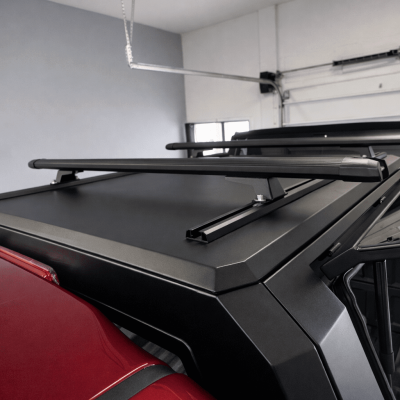 Close-up overhead view of a matte black truck cap roof with integrated roof rack crossbars mounted on a red pickup truck, photographed in a garage.