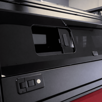 Close-up detail of a black truck cap side panel showing a sliding tinted window and a keyed locking latch mechanism installed on a red pickup truck.